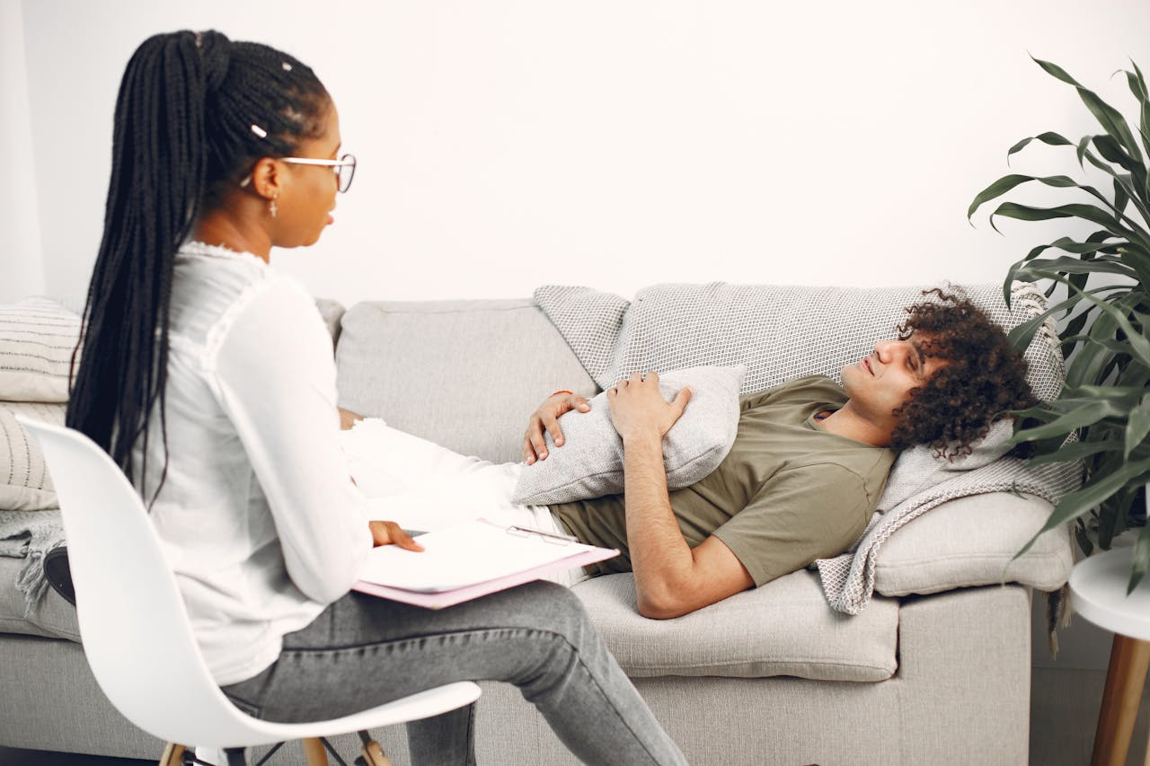 A man in therapy session, lying on a couch, speaking with a female therapist taking notes.