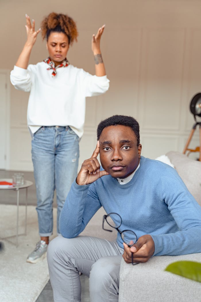 Man sitting pensively, holding eyeglasses, while a woman stands frustrated in the background.